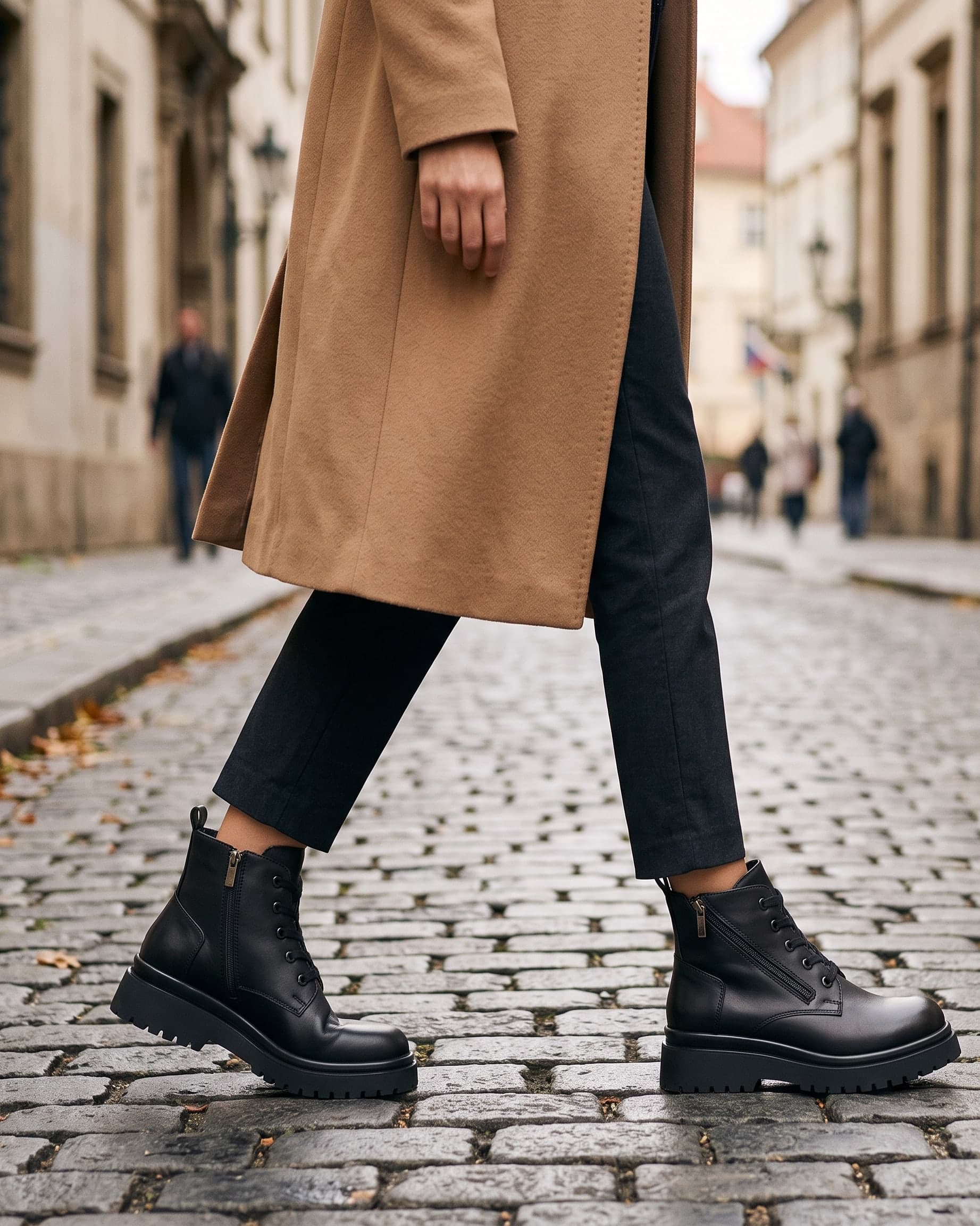 A woman in a camel coat walking down a cobbled street in black leather lace-up ankle boots